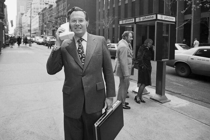 A man in a suit holds an early mobile phone on a city street, representing historical inventions.