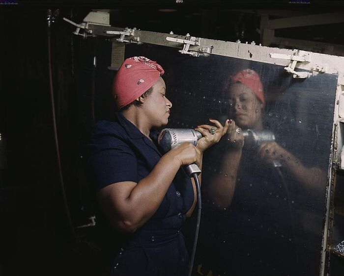 Woman working with a drill, wearing a red headscarf, in a 1940 color photo, reflecting on a metal surface.