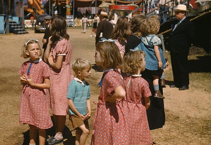 Children in colorful 1940s clothing at a fairground, surrounded by rides and onlookers.