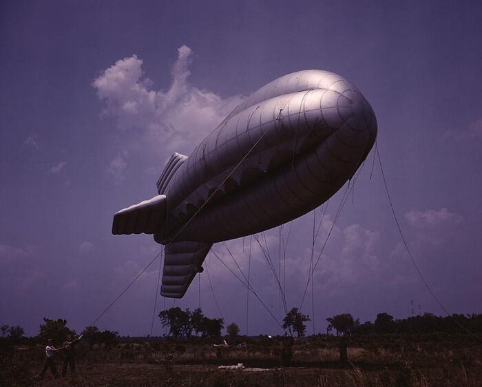 Large silver blimp anchored to the ground in the countryside, under a vivid blue sky, from 1940 color photos.