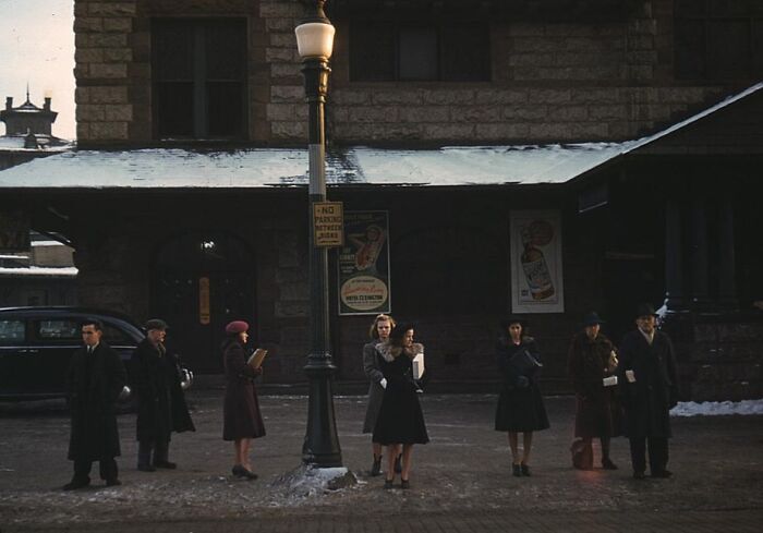 People wait at a snowy street with a vintage car and building in the background, reminiscent of 1940 color photos.