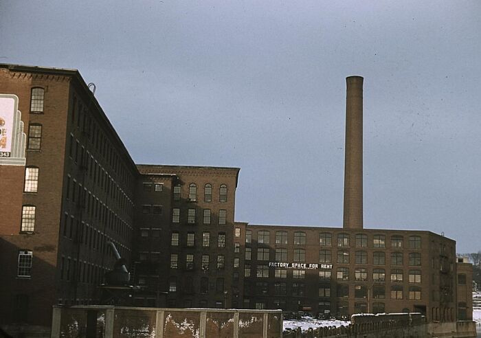 1940 color photo of a vintage brick factory building with a tall smokestack under a cloudy sky.