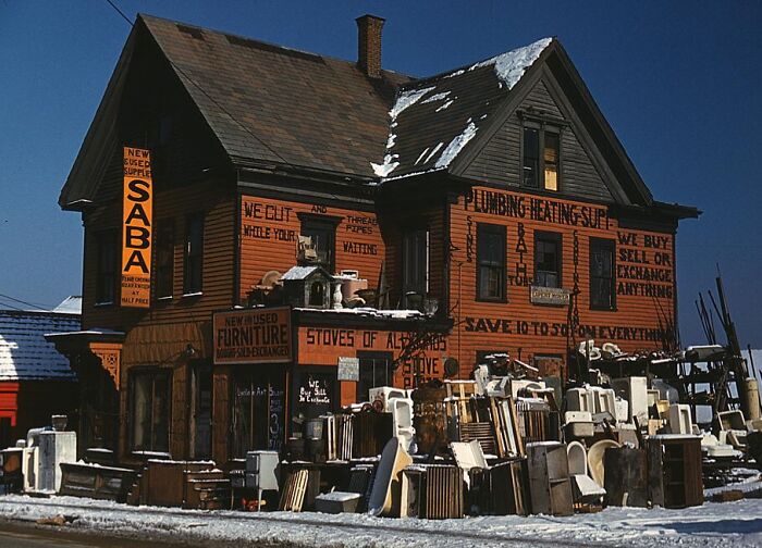 1940 color photo of a vintage store with signs and assorted furniture outside, surrounded by snow.