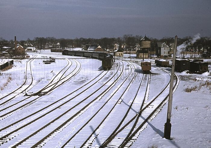 Snow-covered railway tracks in a 1940 color photo, with buildings and a train visible in the distance.