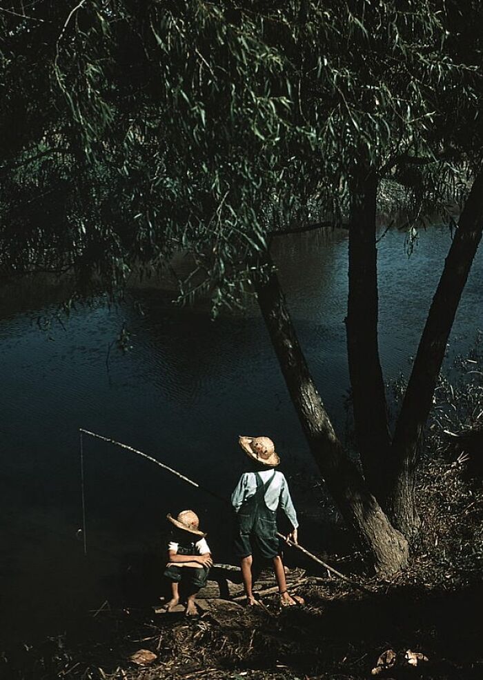 Two children fishing by a river in 1940, wearing hats and overalls, under a tree in a color photo.