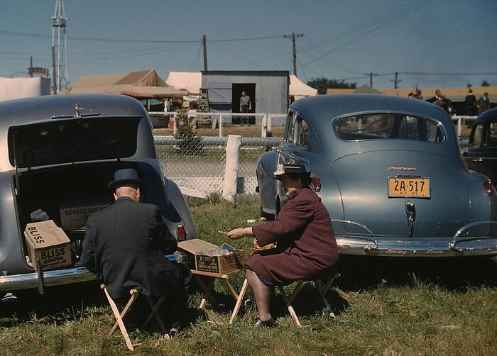 Couple enjoying a picnic by their cars at a fairground in 1940, showcasing vibrant colors and vintage style.