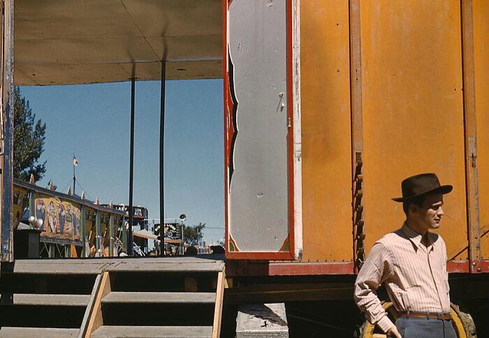 Man in a 1940s color photo stands by a carnival trailer, wearing a hat and striped shirt.