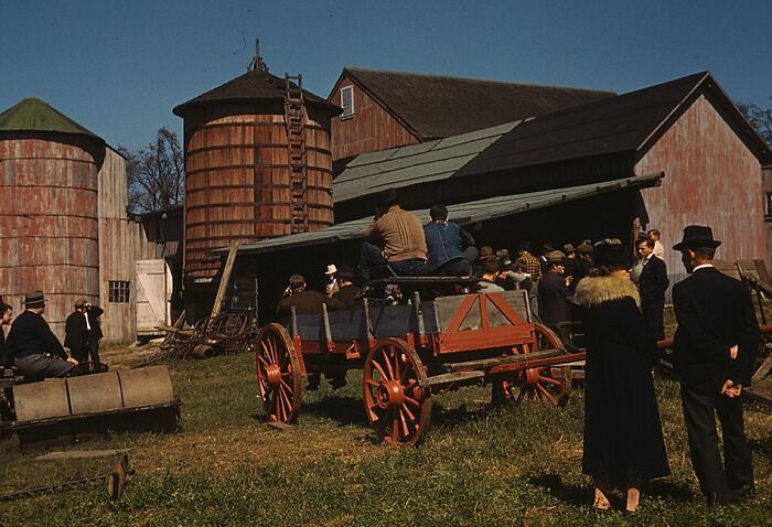 A group gathered around vintage farm buildings with a wagon, capturing a scene from the 1940s in color photos.