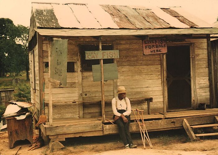 Man sitting on porch of an old wooden building with "live fish for sale" signs, 1940 color photo.