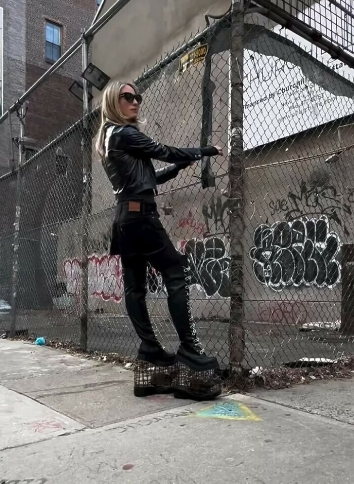 NYC local in edgy fashion, wearing a black leather jacket and platform boots near a graffiti-covered fence.