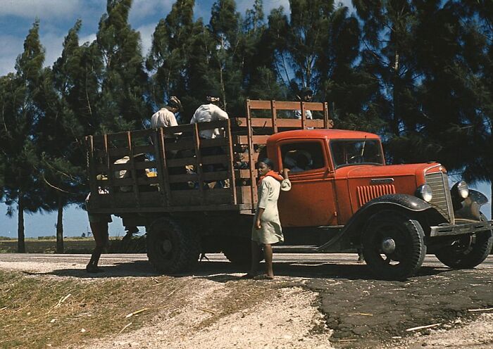 1940 color photo of a red vintage truck with wooden sides, parked on a rural road, with people and trees in the background.