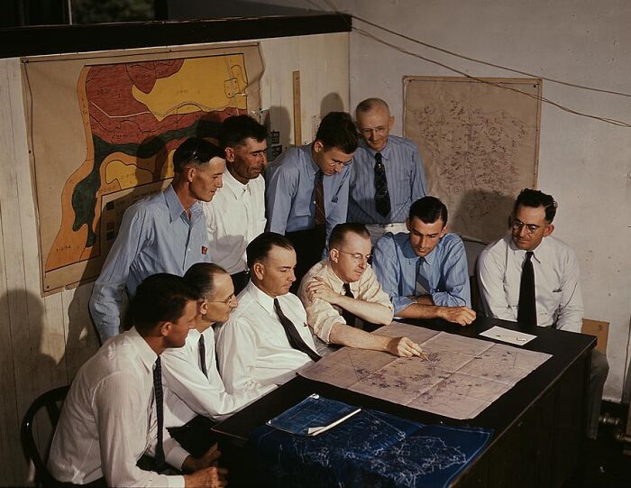 Group of men in 1940 color photo, gathered at a table, analyzing a map with charts on the wall in the background.