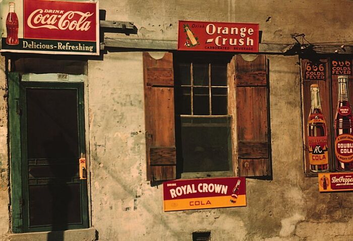 Vintage 1940 color photo with Coca-Cola, Orange Crush, and Royal Crown Cola signs on a weathered building wall.