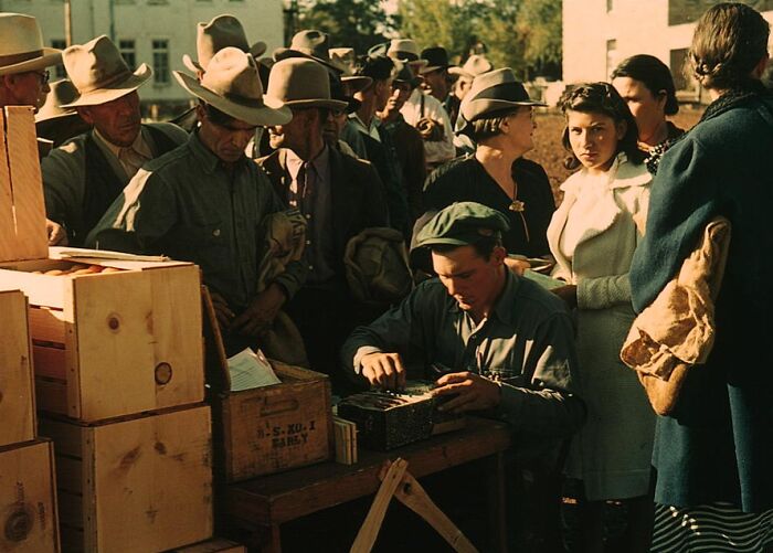 People in 1940 color photo gathered around a man seated at a table with boxes, wearing vintage hats and coats.