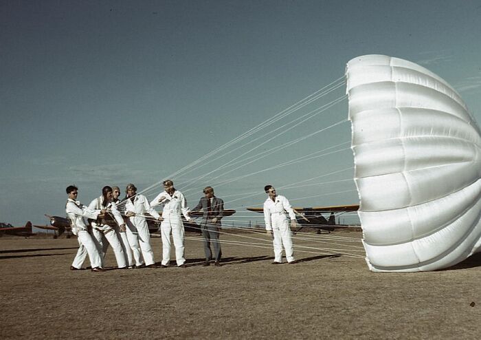 Men in white uniforms holding onto a parachute, vintage 1940 color photo showing aviation training on a grassy field.