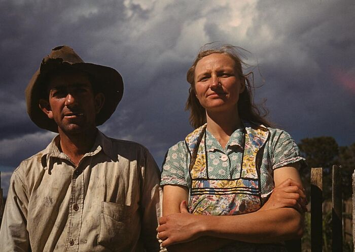 Man and woman outdoors in 1940 color photo, wearing rustic clothing, with a dramatic cloudy sky in the background.