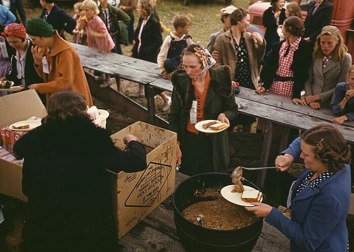 People in 1940s attire at an outdoor food line, receiving plates of stew.