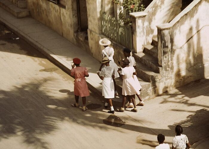 Group of women in 1940 color photo walking down a street beside a building, wearing vintage dresses and hats.