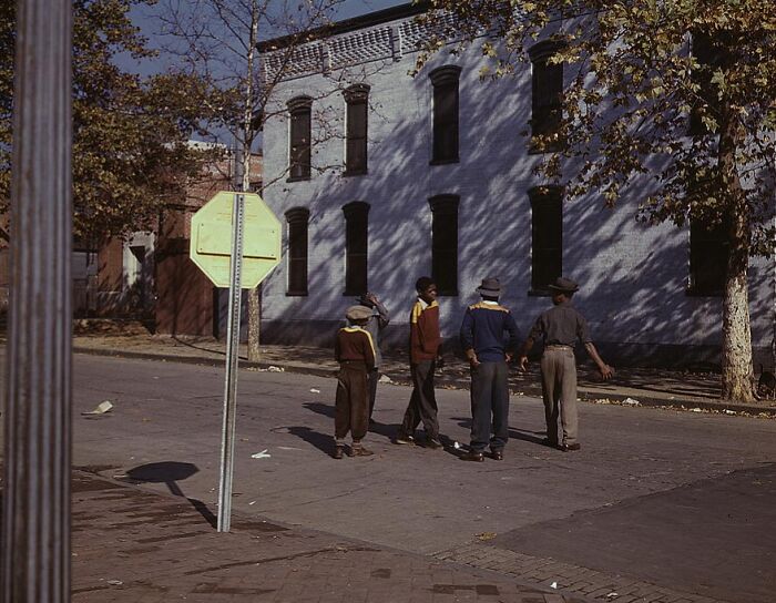 Four people walking on a 1940 street with trees and a two-story building in the background, captured in color.