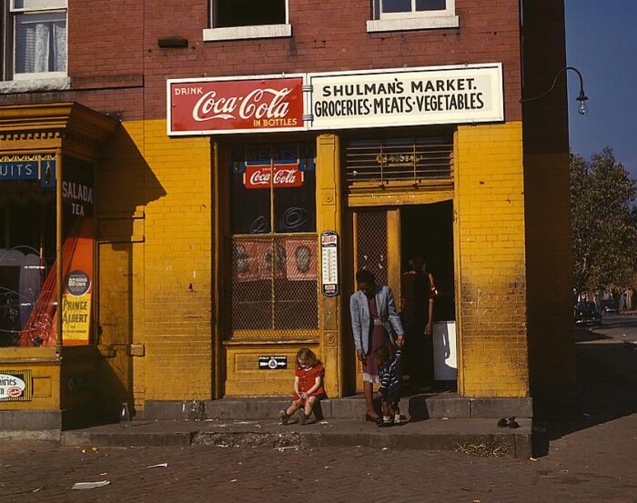 1940 color photo of people outside Shulman’s Market with Coca-Cola sign, showcasing a vintage yellow brick storefront.