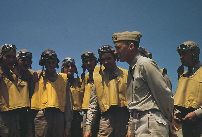 Pilots in 1940 color photo, wearing aviator gear and life vests, gathered under a clear blue sky.