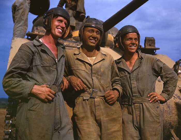 Three soldiers in 1940s color photo, smiling in front of a tank, wearing helmets and coveralls.