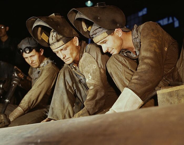 "Workers wearing welding helmets in a 1940 color photo, focused on a project in a dimly lit environment."