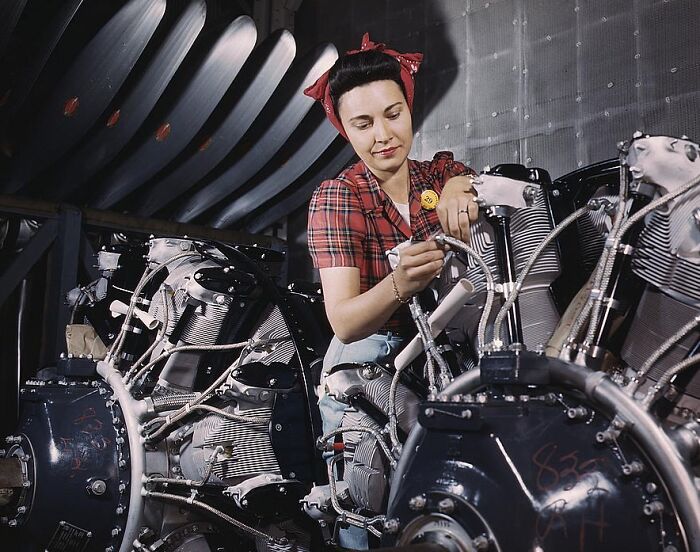 Woman in 1940s attire working on aircraft engines in a colorful photograph.