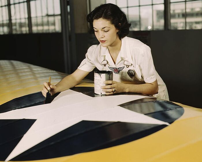 Woman painting aircraft insignia in a hangar, 1940s color photo.