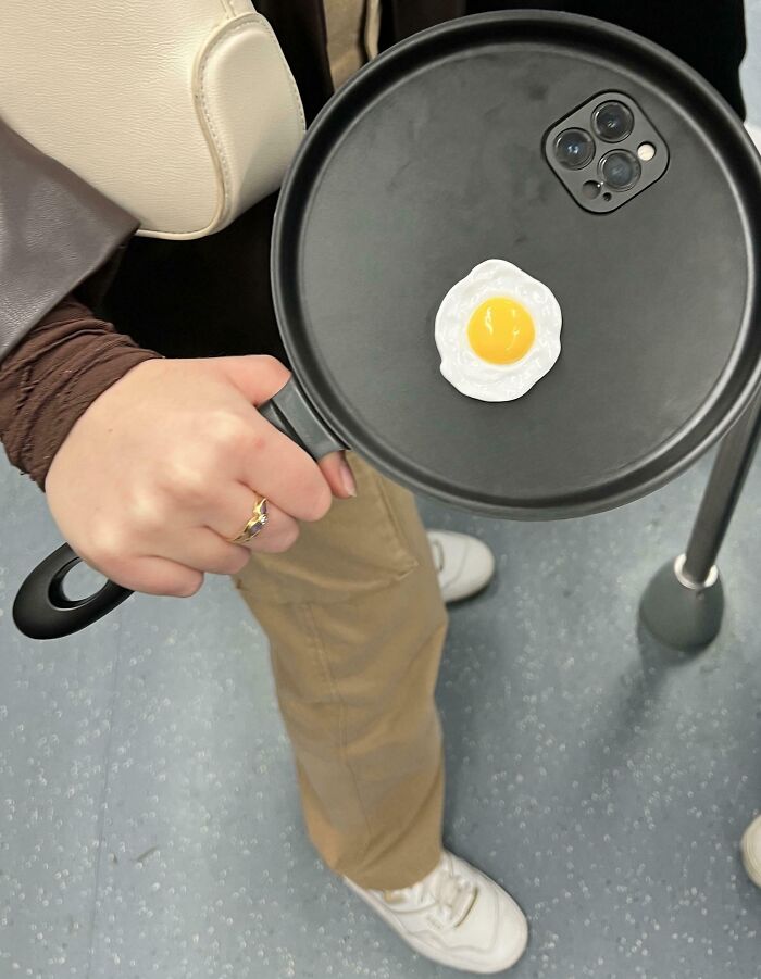 Subway passenger holding a frying pan with a phone inside, next to a toy egg, creating a humorous scene.