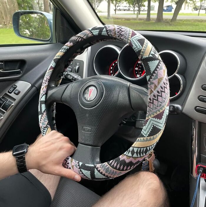 Person holding a steering wheel with a colorful patterned cover in a car interior, showcasing unique car items.