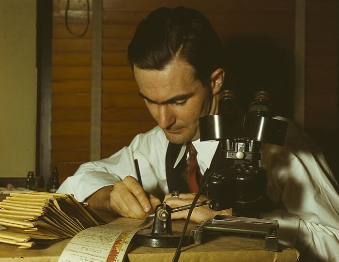 Man in 1940 using a microscope, wearing a suit and tie, takes notes at a desk with stacks of papers.