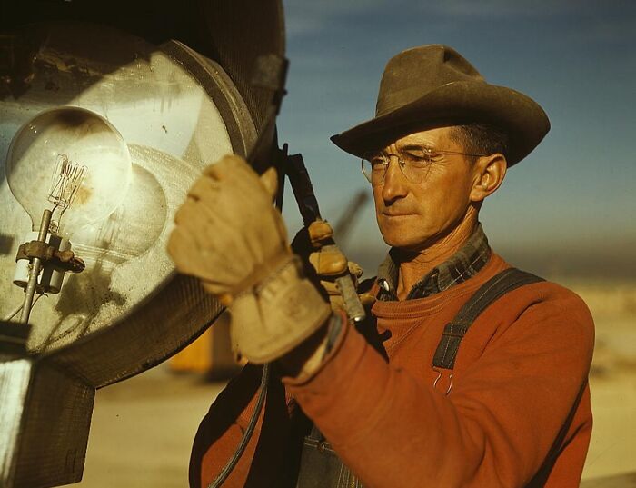 Man in 1940s attire, wearing glasses and a hat, adjusting a large vintage light fixture outdoors, in a color photo.