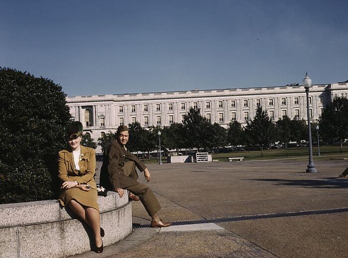 Two people in 1940s attire sit outdoors, with a large building in the background.