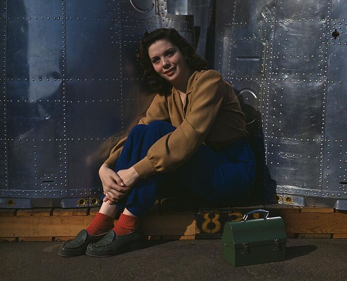 Woman in 1940s clothing sitting by a metal structure, with a green lunchbox on the ground.