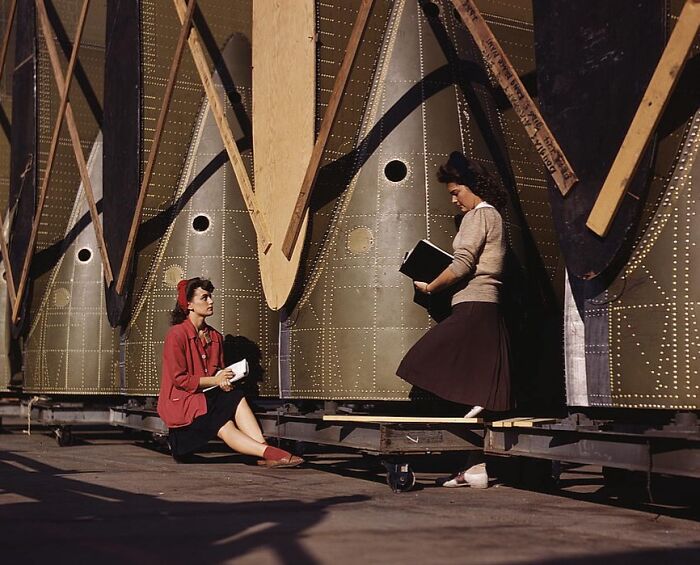 Women working on aircraft components, wearing 1940s attire, with large metal structures in the background.