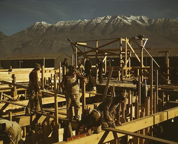 Construction workers building wooden structures in a 1940 color photo with mountains in the background.