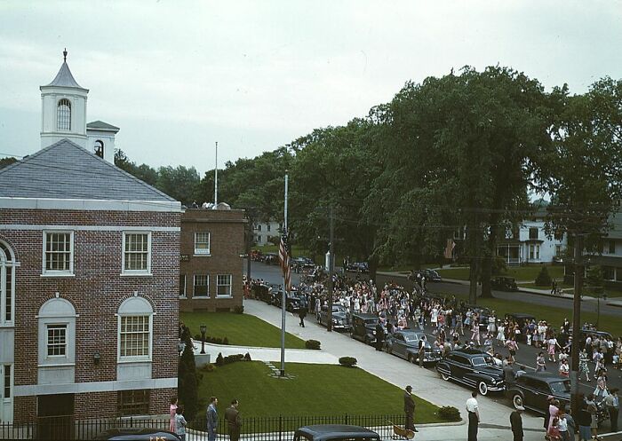 1940 color photo of a parade with vintage cars and crowds beside brick buildings and lush trees.