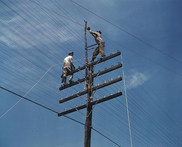 Workers on a telephone pole in a 1940 color photo, set against a clear blue sky.