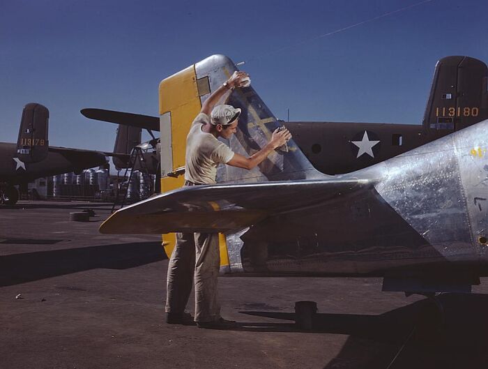 Worker polishing aircraft tail, 1940 color photos, on an airfield under a clear blue sky.