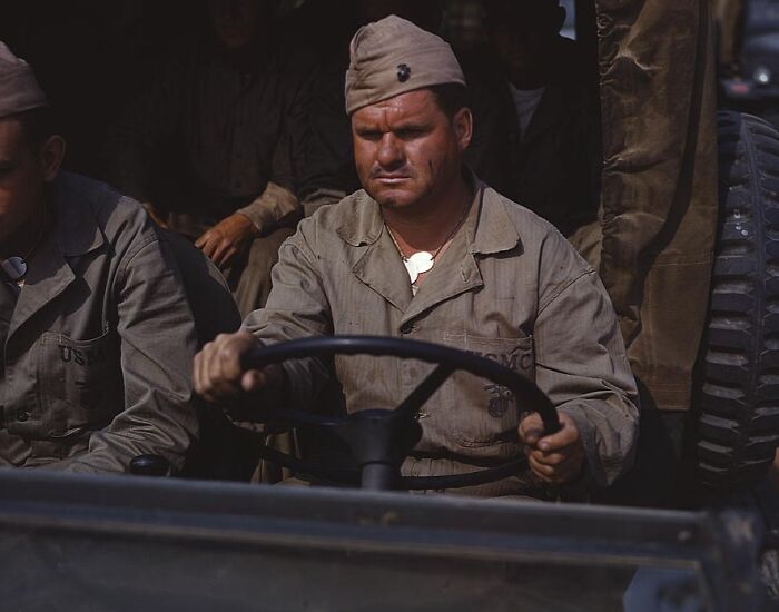 Soldier driving a military vehicle in 1940, wearing a uniform, with serious expression and a comrade seated beside him.