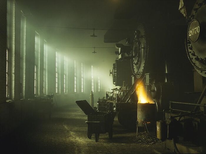 Historic 1940 color photo of a dimly lit factory interior with machinery and a glowing furnace.