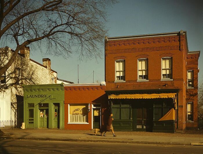 1940 color photo of a street with vintage red brick building, green laundry, and bakery storefronts under a clear sky.