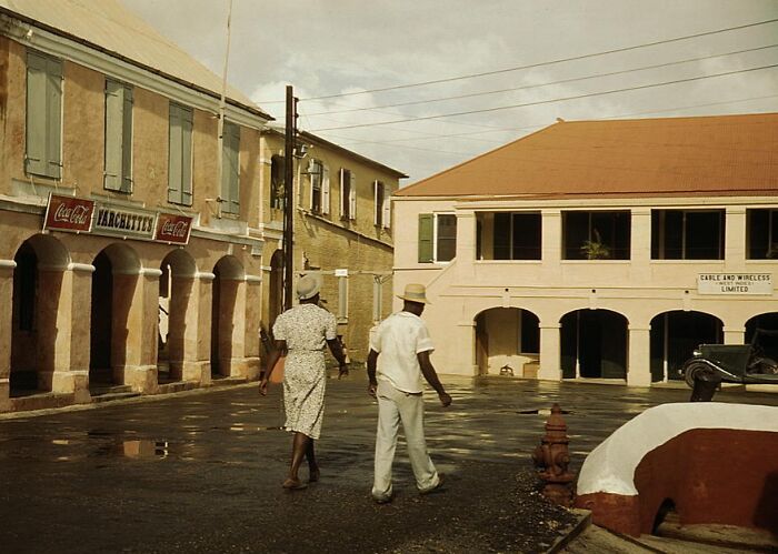 Historic street scene with two people walking past 1940 color photos of vintage buildings and storefronts.