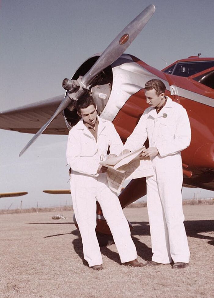 Two men in white jumpsuits reviewing a document in front of a plane, captured in 1940 color photos.