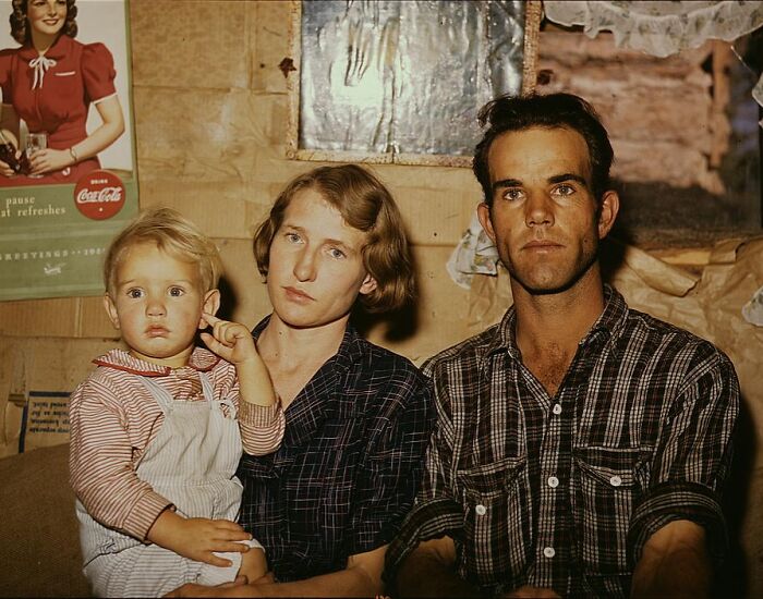 Family sitting together in 1940 color photo, showing a man, woman, and child inside a rustic home with a vintage poster.