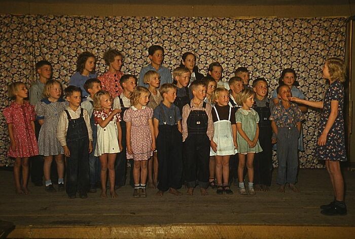 Children in a 1940s choir, wearing colorful clothes, performing on a stage with a floral backdrop.