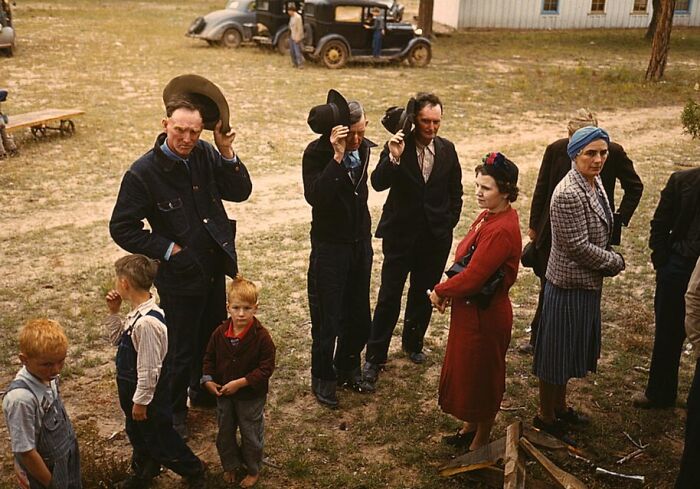 People in vintage attire during a 1940s outdoor gathering, with vintage cars and period fashion in the background.