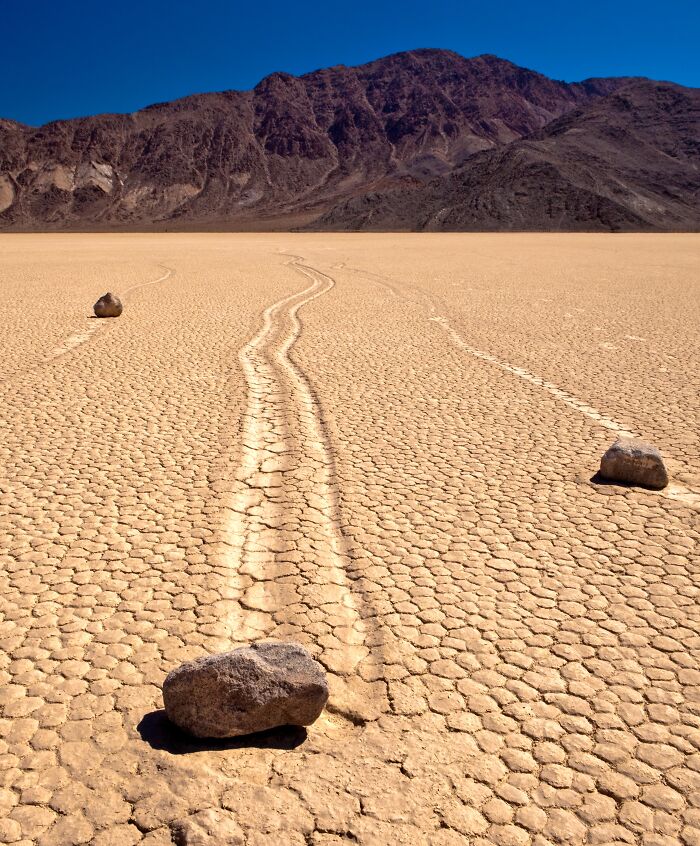 Rocks create tracks on sun-cracked desert floor, showcasing strange natural phenomena under a clear blue sky.