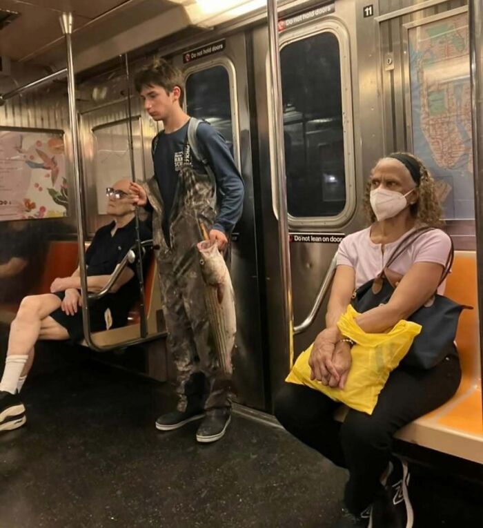 Man in overalls holding a large fish on a subway train with other funny passengers seated nearby.